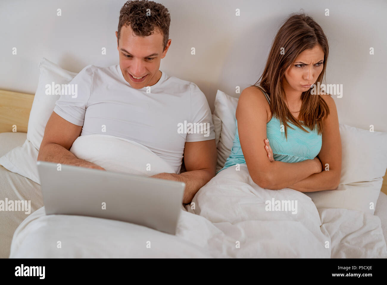 Giovane con il computer portatile a letto. Soddisfatto l uomo che navigano in rete al computer portatile, donna sconvolto e arrabbiato guardando l'altro lato. Foto Stock