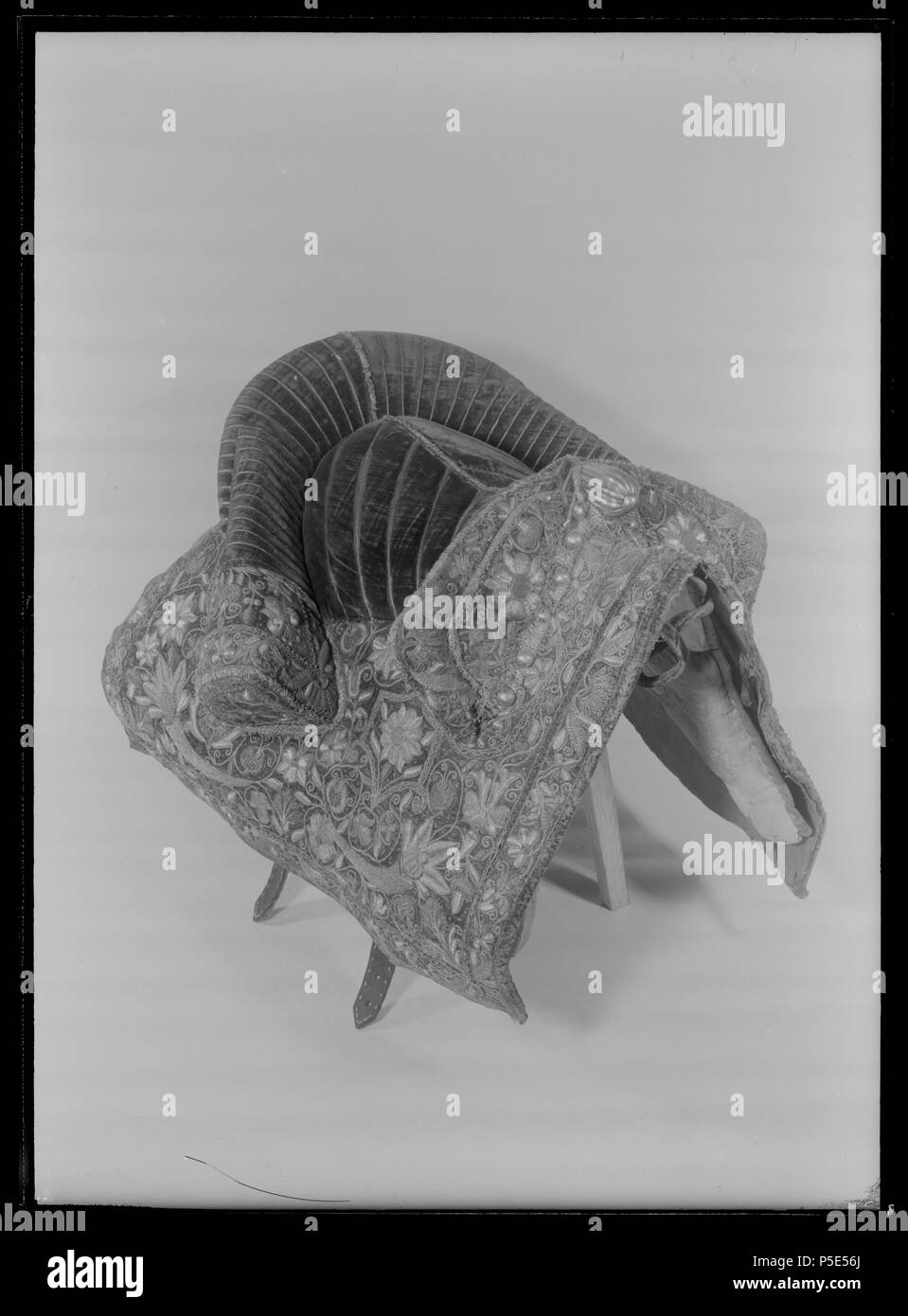 Bomsadel . Svenska: Överklädd med purpurröd sammet. Bakbommen med kraftiga stödbommar. Sätet nersytt med smala flätband ho ett glest fiskbensmönster. De båda bommarnas insidor med vertikalt påsydda, sådana band. Bommarnas utsidor samt sadeltäcket har rika sollievo och läggsömsbroderier ho guldtråd, stora blommor, ekollon, palmetter och ymnighetshorn. Utmed bommarnas utsidor fanns ursprungligen tännlikor av mässing förgylld, numera återstår endast 12 utmed den främre bommen. Framåtriktad, profilerad sadelknapp av mässing förgylld. Sadeltäcket fodrat med ljust läder. Inre Sätets längd: 320 mm.Bakbo Foto Stock