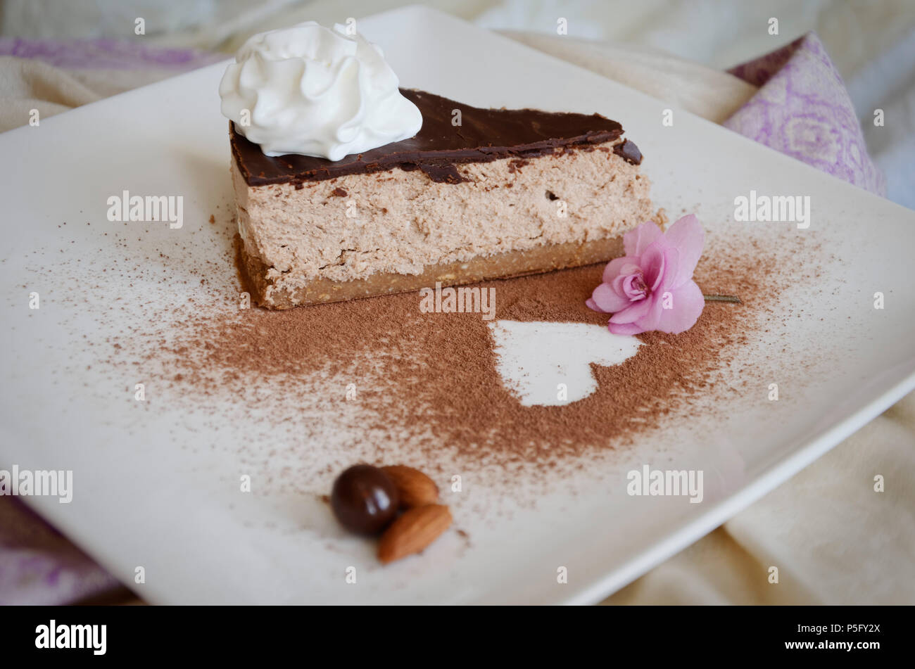 Pezzo di torta al cioccolato con panna montata sulla parte superiore su una lastra decorata con fiore violaceo, mandorle e cosparsi di cioccolato in polvere. Foto Stock