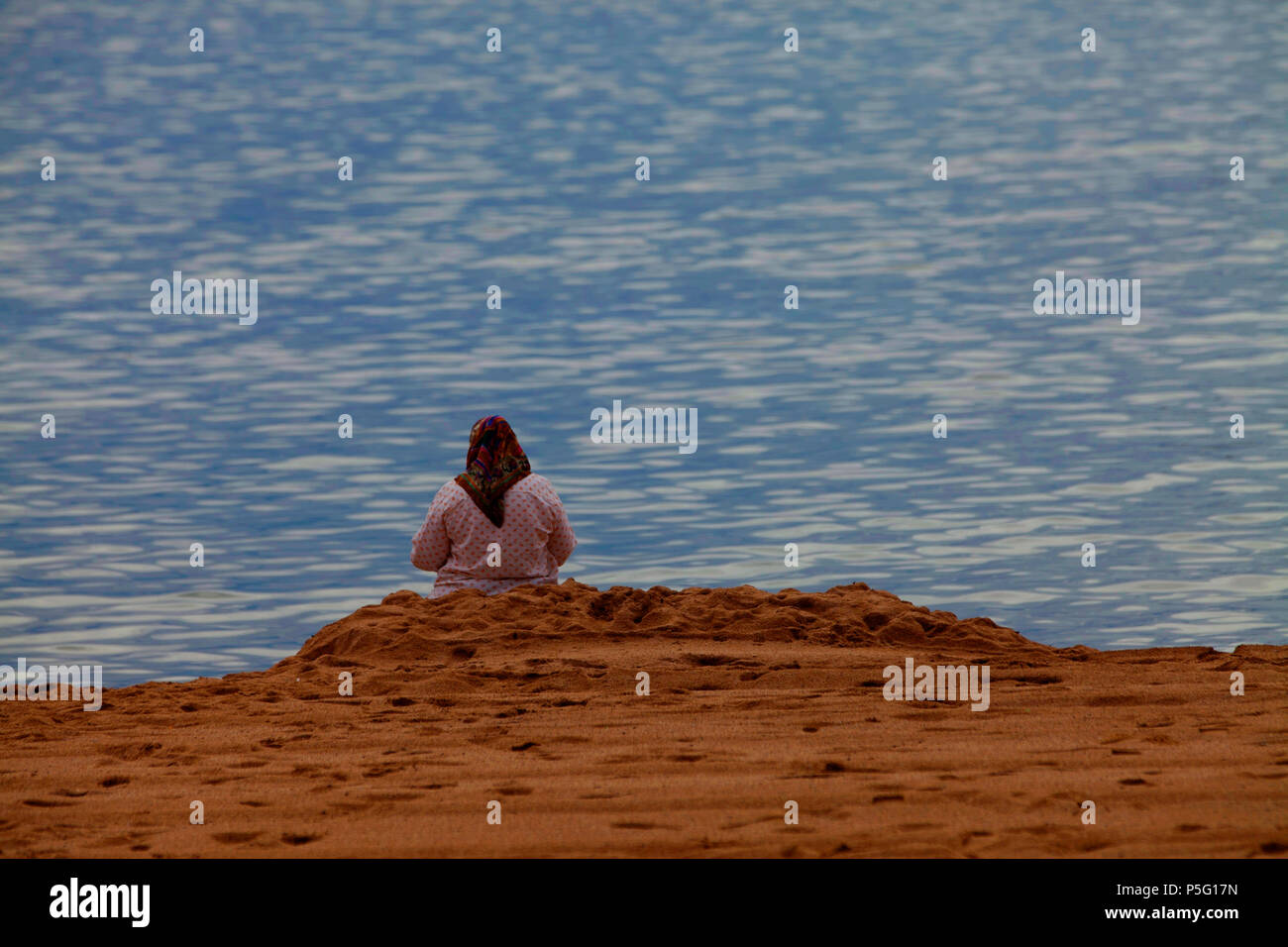 Eine Frau mit Kopftuch von hinten die alleine am Strand sitzt und auf das Meer schaut in Cannes, Frankreich. Foto Stock