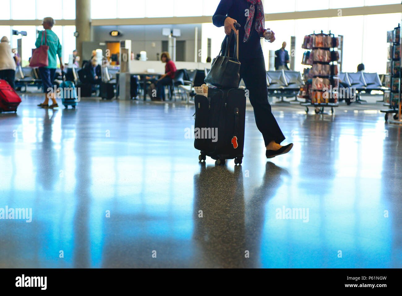 Una vista di persone, una donna dal collo in giù, rendendo il loro modo attraverso il terminal di un aeroporto con piani riflettenti, tirando bagagli da e per il Foto Stock