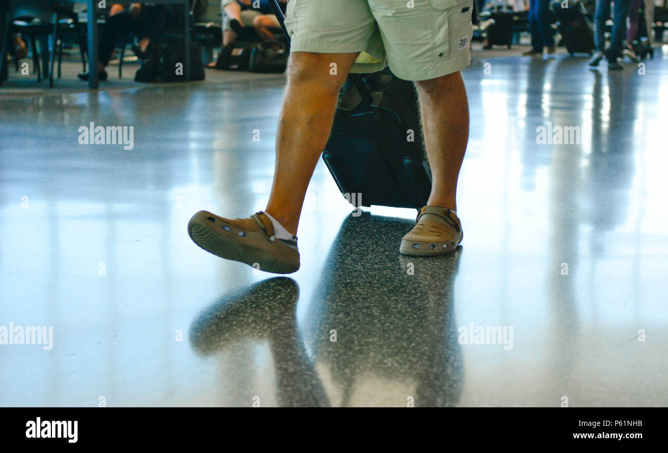 Una vista dal basso della vita di un uomo in pantaloncini corti che fa il suo modo attraverso il terminal di un aeroporto con piani riflettenti, rullo di trascinamento per i bagagli o da un Foto Stock