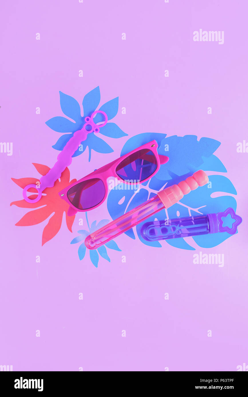 Bacchette di bolla e occhiali da sole su un pastello sfondo viola con foglie tropicali e copia di spazio. Attività estive il concetto di overhead Foto Stock