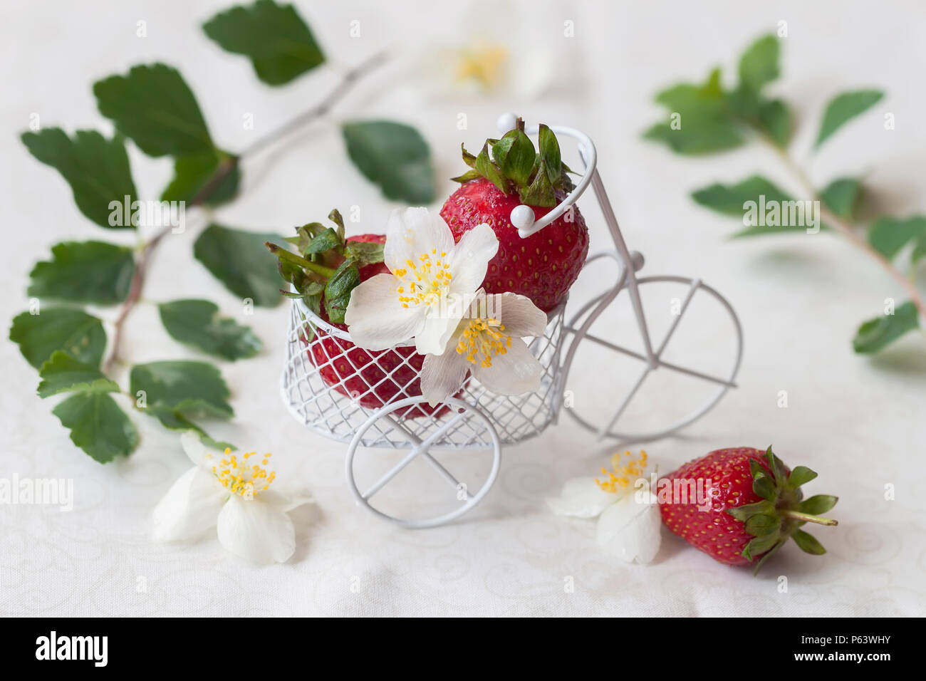 Giocattolo in miniatura Carrello Il carrello closeup con fragole mature, foglie, fiori su sfondo chiaro. Concetto di cibo sano, detox, vitamine, dieta Foto Stock