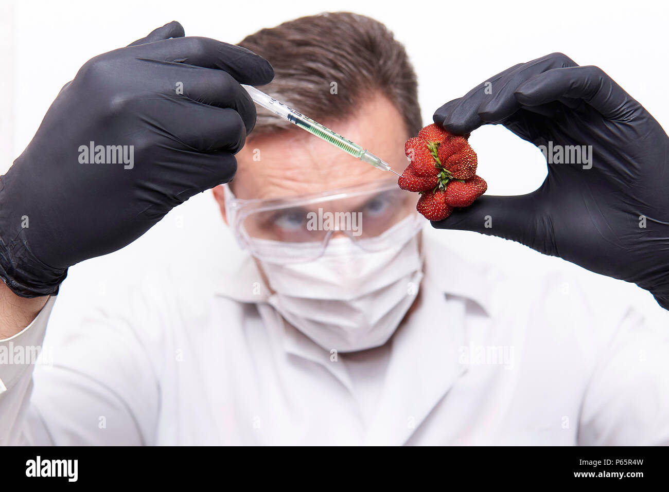In una fragola di forma insolita, uno scienziato in occhiali protettivi, una maschera e guanti di protezione rende una iniezione su uno sfondo bianco. Foto Stock