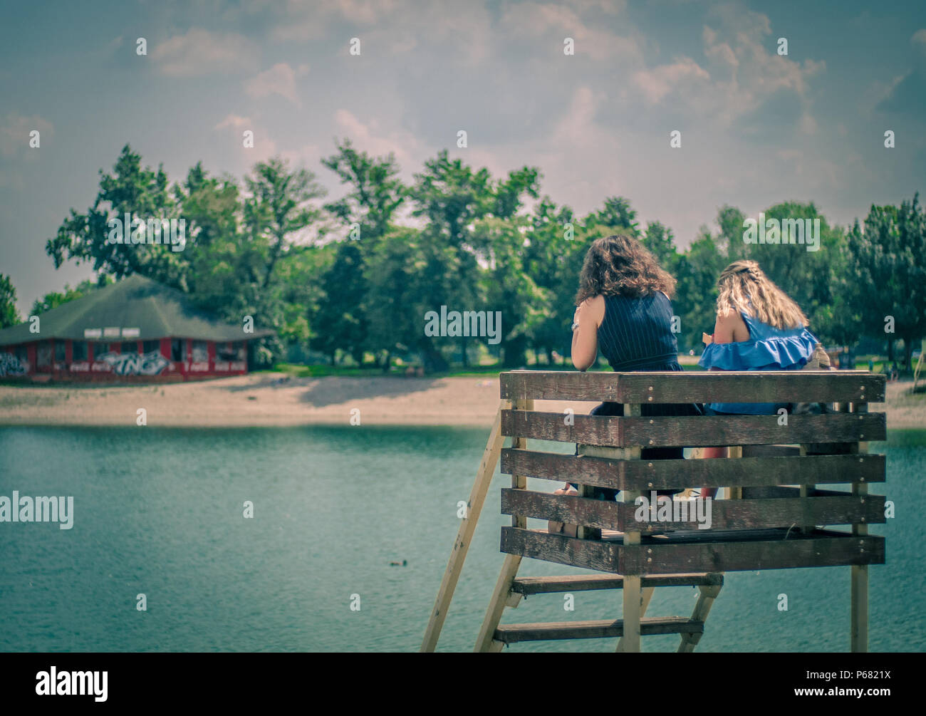 Vista posteriore di due ragazze adolescenti seduti su un alto seggio presso la riva del lago, godendovi la vista su un caldo giorno d'estate. Foto Stock