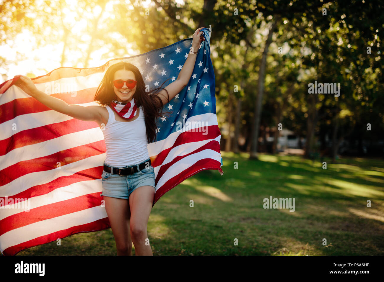 Ragazza in pantaloncini corti in esecuzione nel parco azienda bandiera americana. Ragazza sorridente con bandiera americana al parco sulla quarta di luglio. Foto Stock