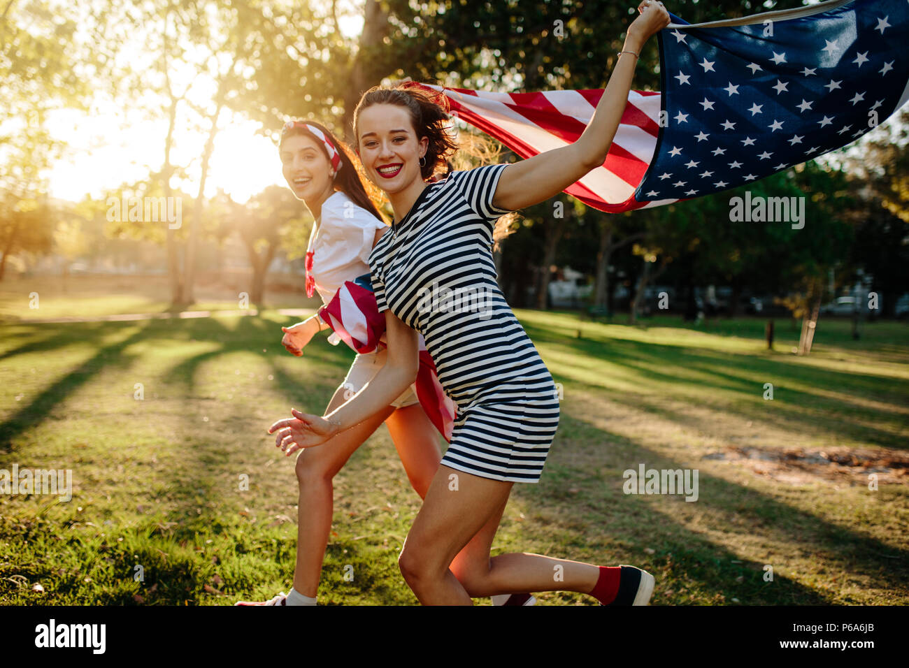 Bella giovane donna con la femmina amico in esecuzione nel parco azienda bandiera americana. Due ragazze con la bandiera degli Stati Uniti godendo all'esterno. Foto Stock