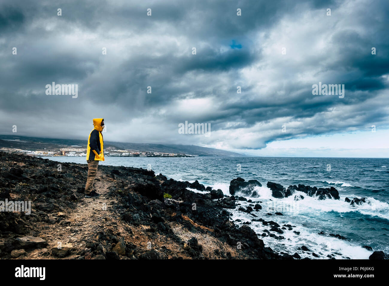 Uomo con giacca gialla in un luogo selvaggio sulla costa dell'oceano guardando la potenza delle onde in un cattivo tempo. sfida e godersi il viaggio lifestyle Foto Stock