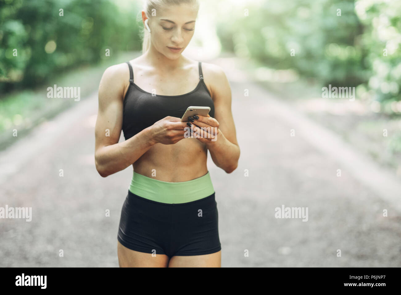 Runner donna nella mattinata estiva Park ascoltare musica sullo smartphone utilizzando gli auricolari Bluetooth. Femmina ragazza Fitness jogging sul percorso esterno. Foto Stock