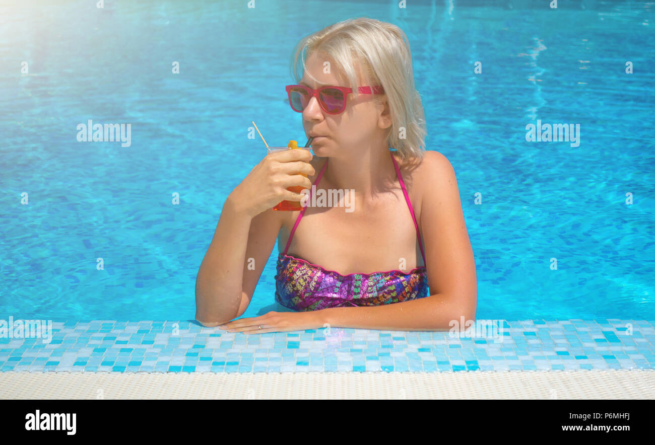 Giovane donna bionda con cocktail drink in piscina. Felice bellissima femmina con eleganti occhiali da sole e bikini bere cocktail e rilassanti in swimmin Foto Stock