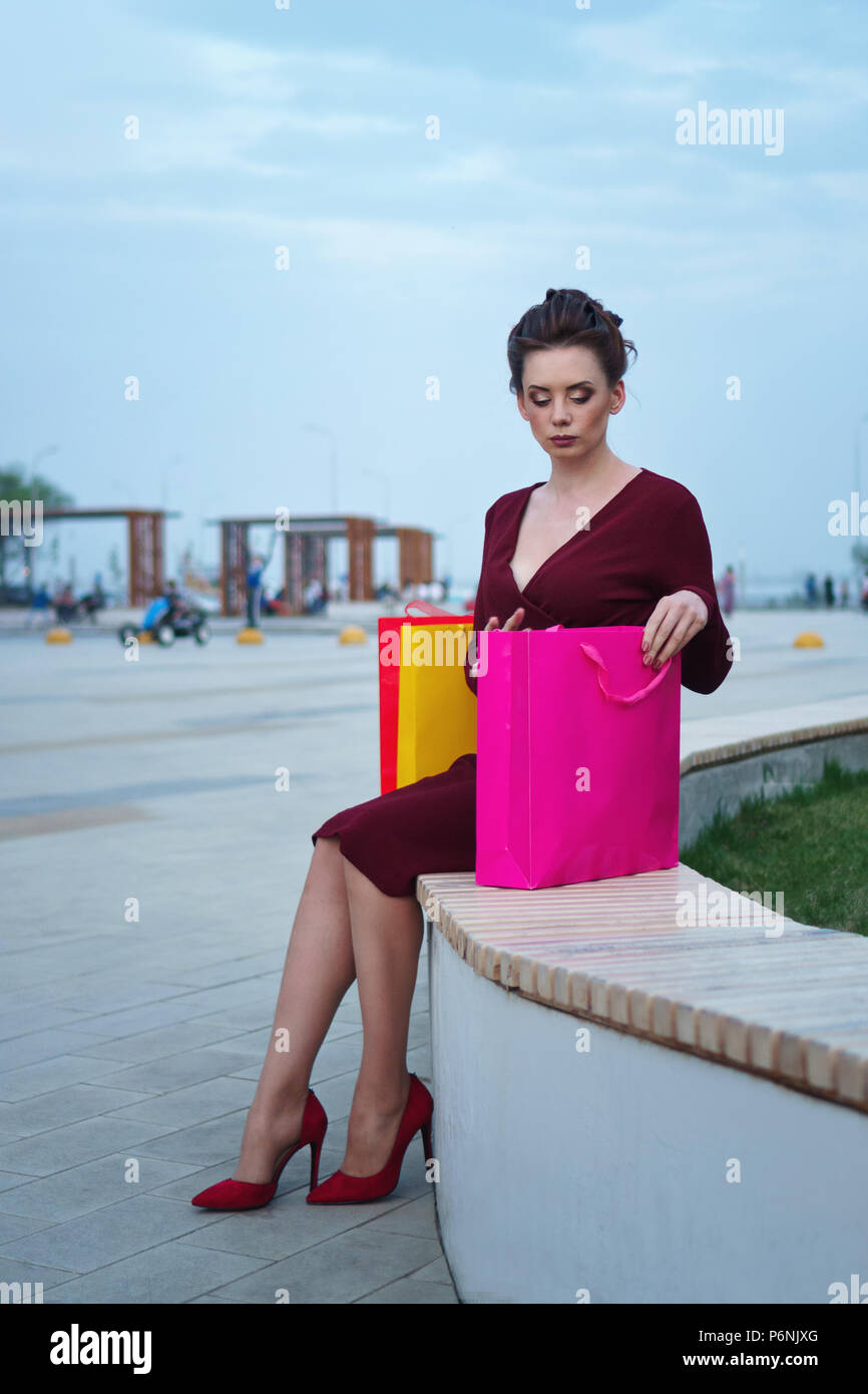 Affascinante ragazza in abito rosso e scarpe con i tacchi alti si siede sul  banco di lavoro. Multicolore di borse per lo shopping sono accanto a lei  Foto stock - Alamy