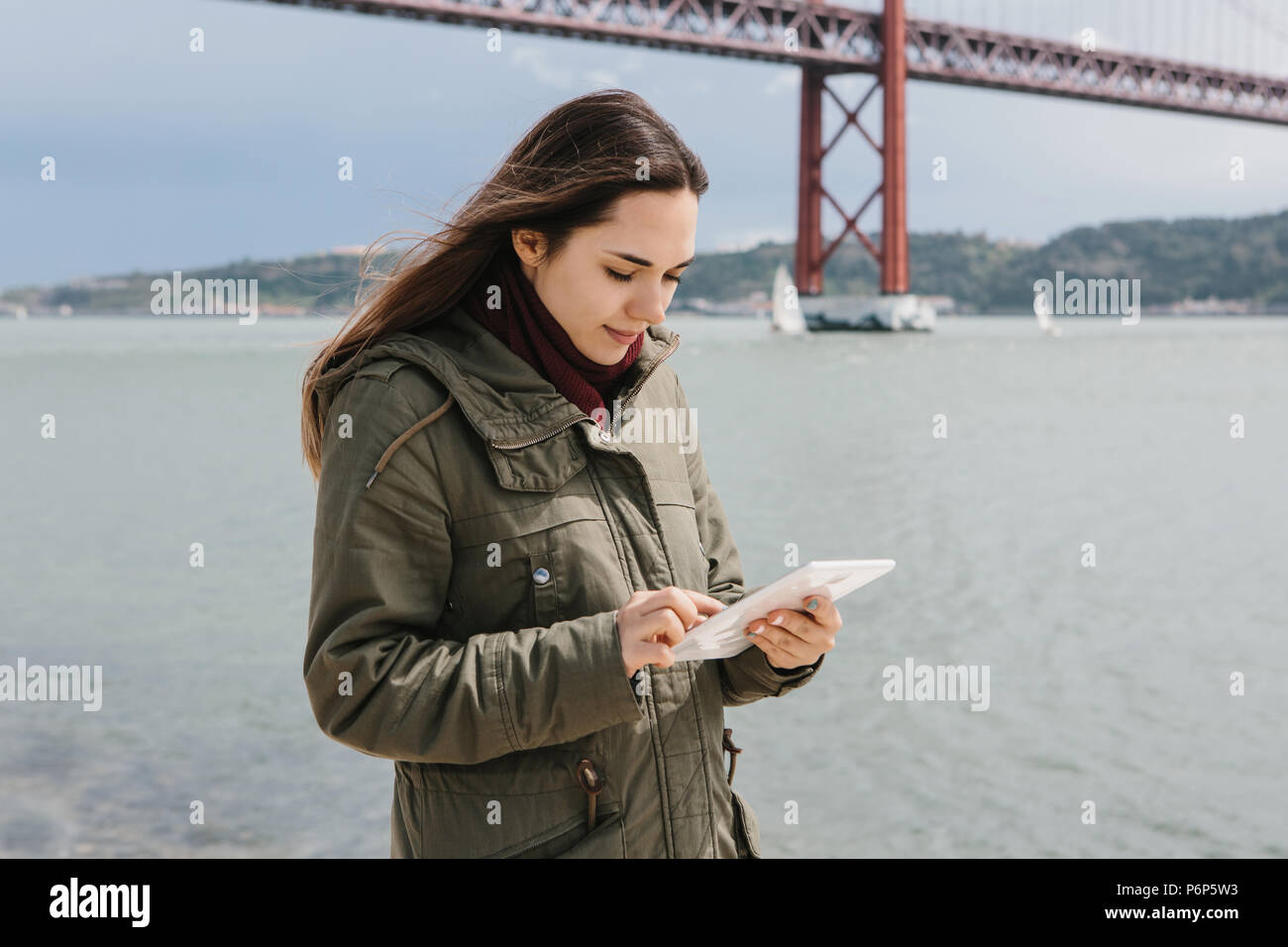 Una giovane donna bellissima utilizza una tavoletta per comunicare con amici o guarda una mappa o qualcos'altro. Ponte 25 Aprile a Lisbona in Portogallo in background. Foto Stock