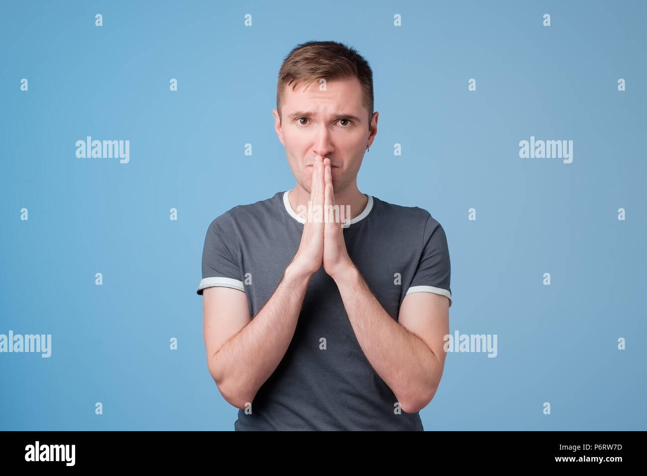 Bel giovane uomo in blu t-shirt pressature palms insieme e guardando la telecamera, elemosinare il perdono Foto Stock