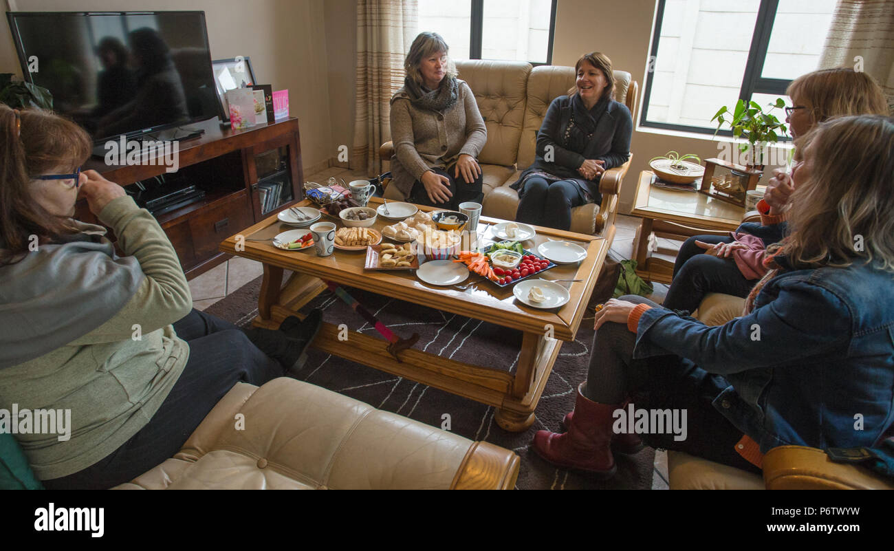 un gruppo informale di donne di mezza età si siede intorno ad un tavolo da caffè a casa piena di una varietà di cibo o spuntini concetto di stile di vita casalingo autentico Foto Stock