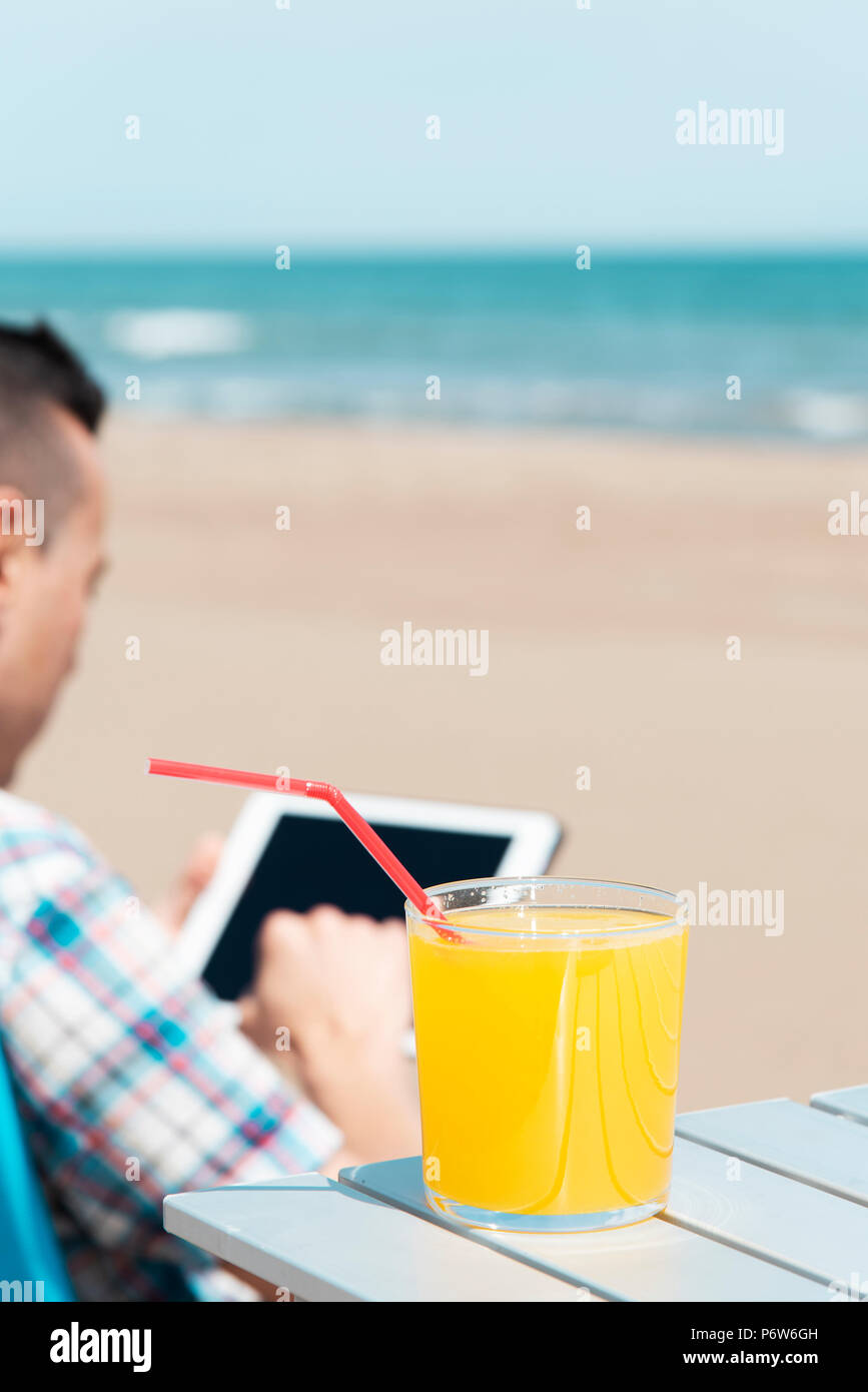 Primo piano di un giovane uomo caucasico sulla spiaggia, utilizzando una tavoletta digitale, seduti su una sedia a sdraio, accanto ad un tavolo all'aperto con un vetro con un refreshi Foto Stock