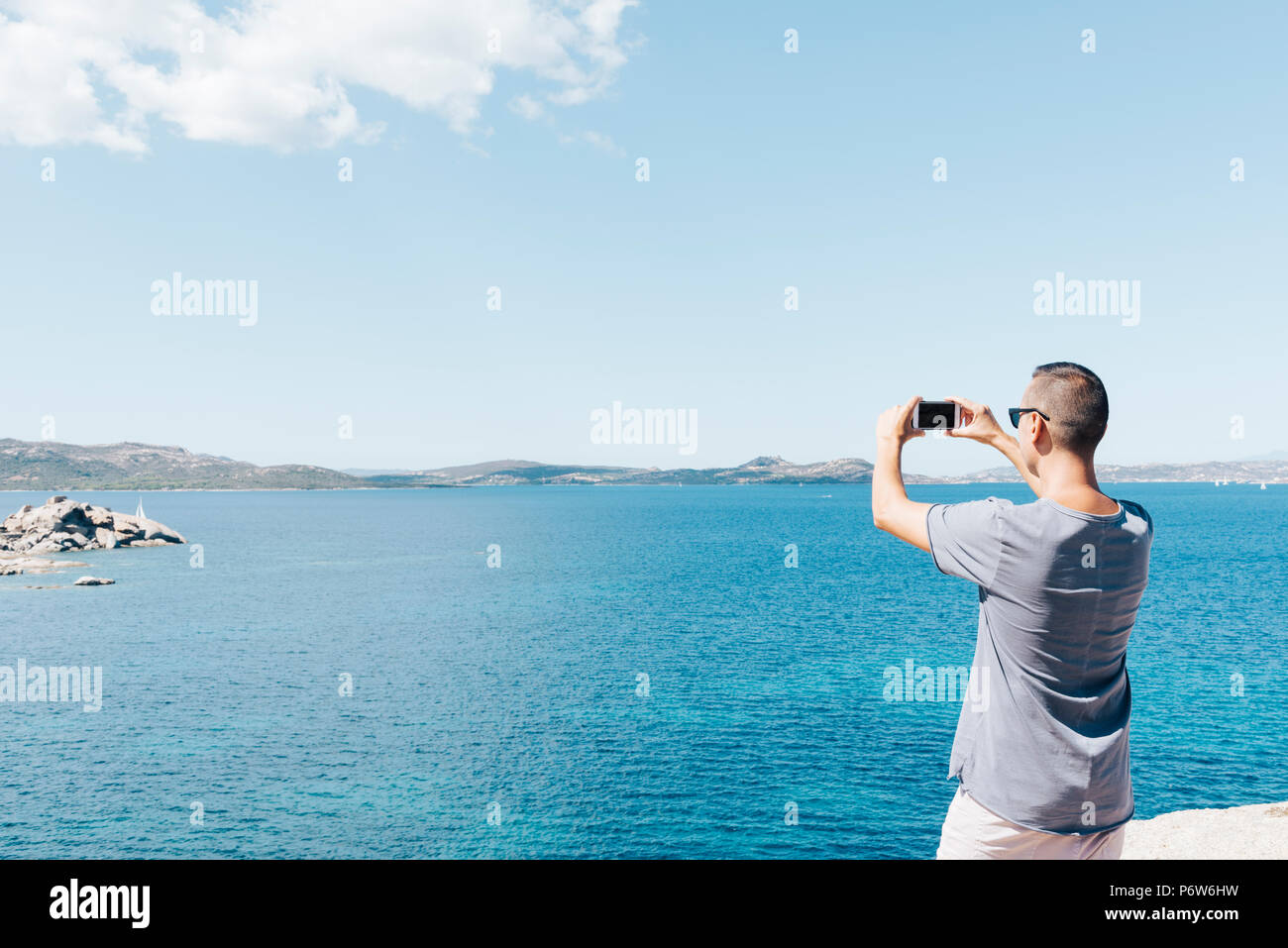 Primo piano di un giovane uomo caucasico visto da dietro di scattare una foto del mare della Costa Smeralda, in Sardegna, Italia, con il suo smartphone, con un bla Foto Stock