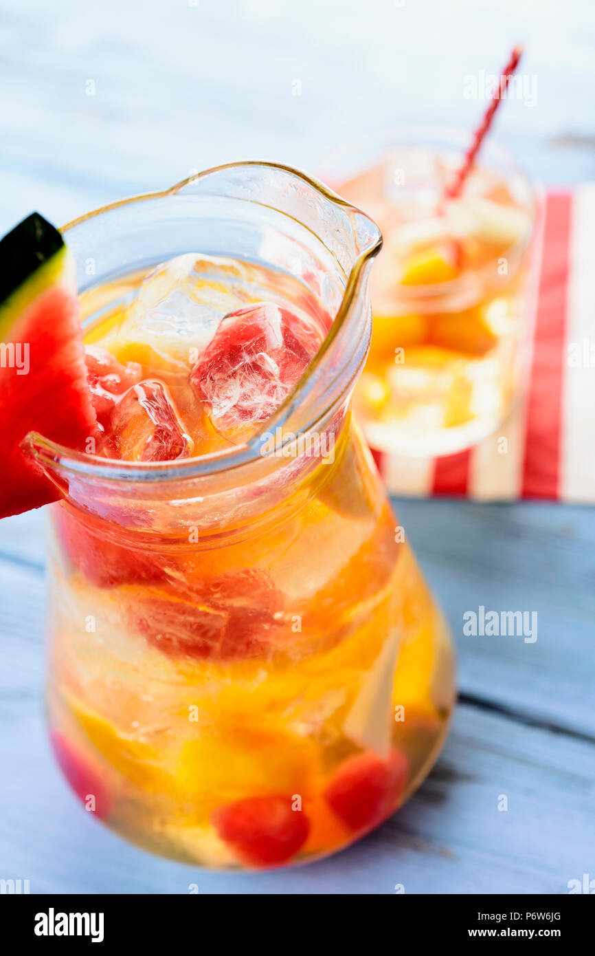 Primo piano di una brocca di vetro con rinfrescanti Sangria spagnola blanca, bianco sangria, con pezzi di frutta fresca, su un blu tavola in legno rustico Foto Stock