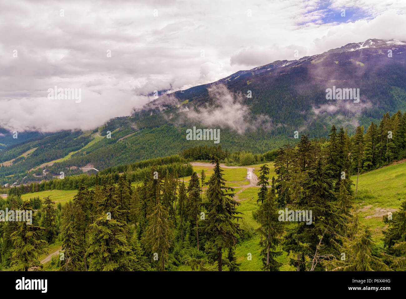 Soffici nuvole bianche al di sopra delle montagne boscose con strade di campagna, alberi di conifere e la neve sulla sommità Foto Stock