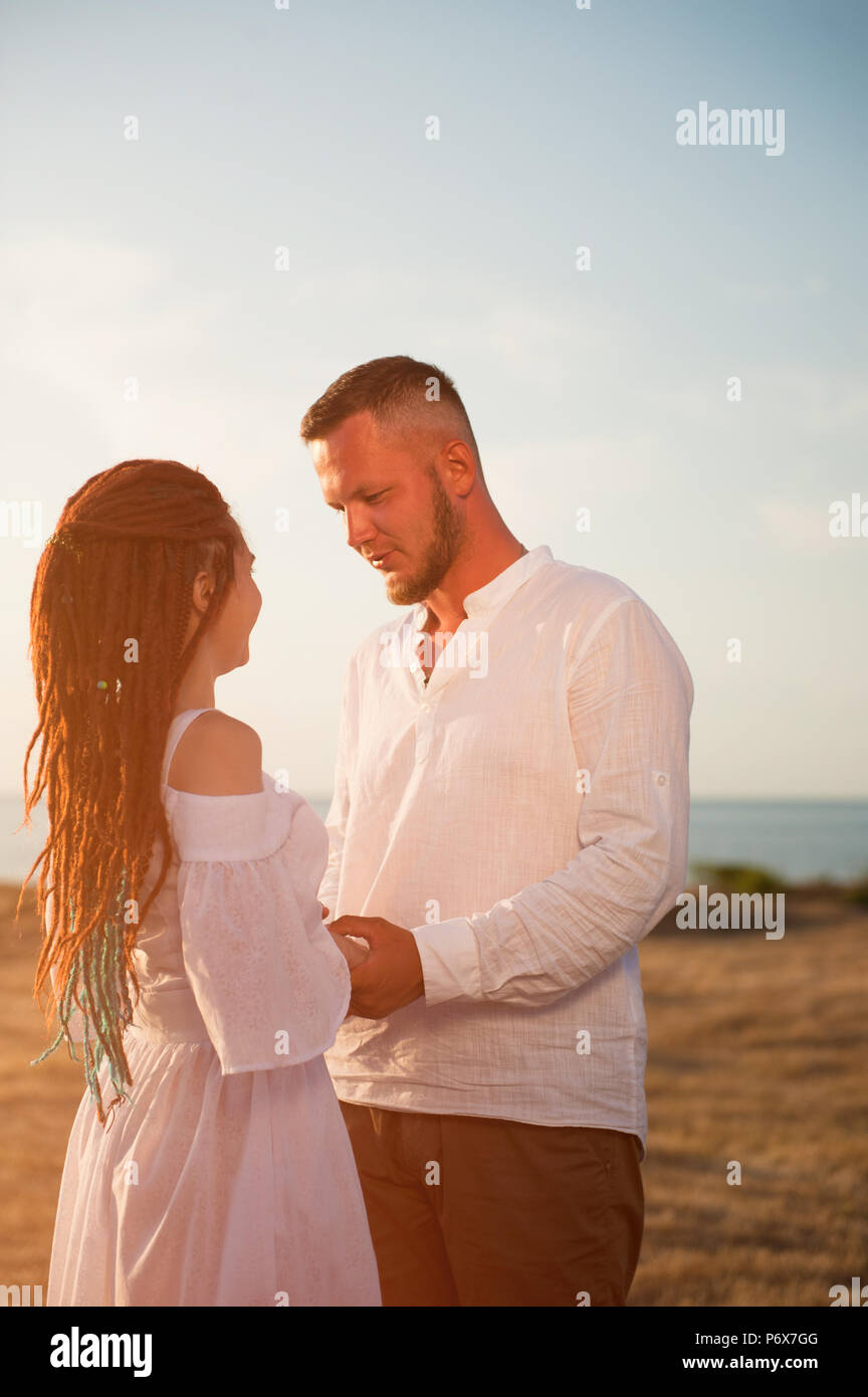 Carino giovane maschio barbuto rende la proposta di matrimonio bella ragazza con dreadlocks in estate tramonto Foto Stock