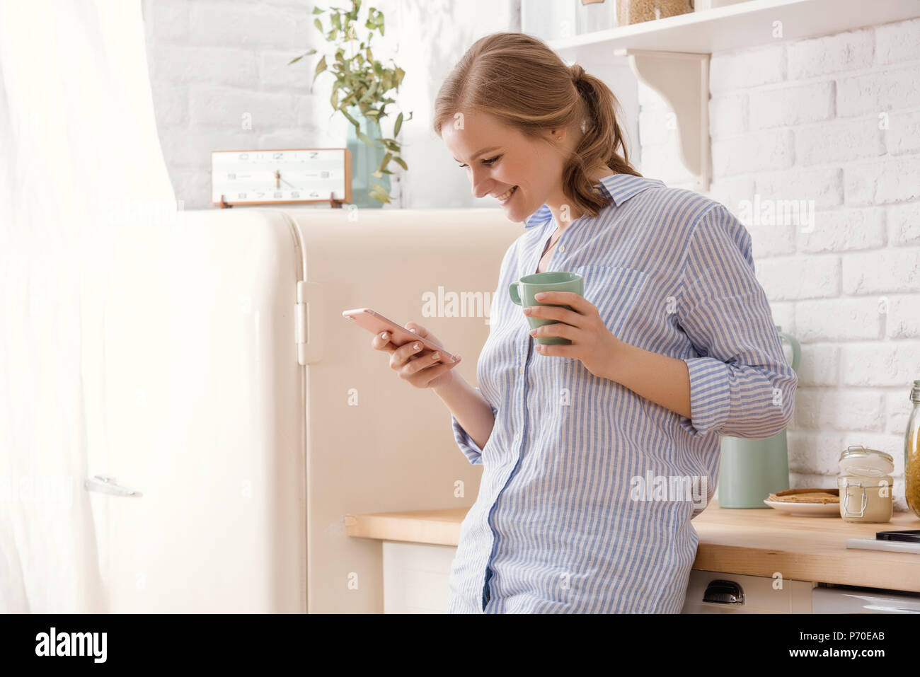 Giovane donna utilizza lo smartphone appoggiata al tavolo della cucina con tazza da caffè e agenda elettronica in una casa moderna. Donna sorridente lettura messaggio telefonico. Bruna ha Foto Stock