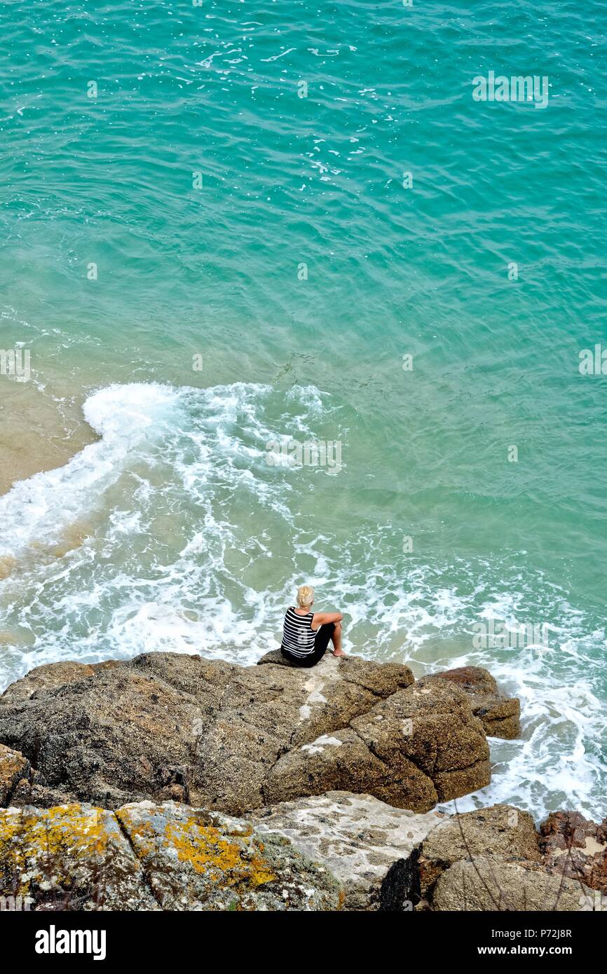 Persone di mezza età bionda donna seduta sulle rocce circondato da un Cornish blue sea Porthcurno Cornwall Inghilterra REGNO UNITO Foto Stock