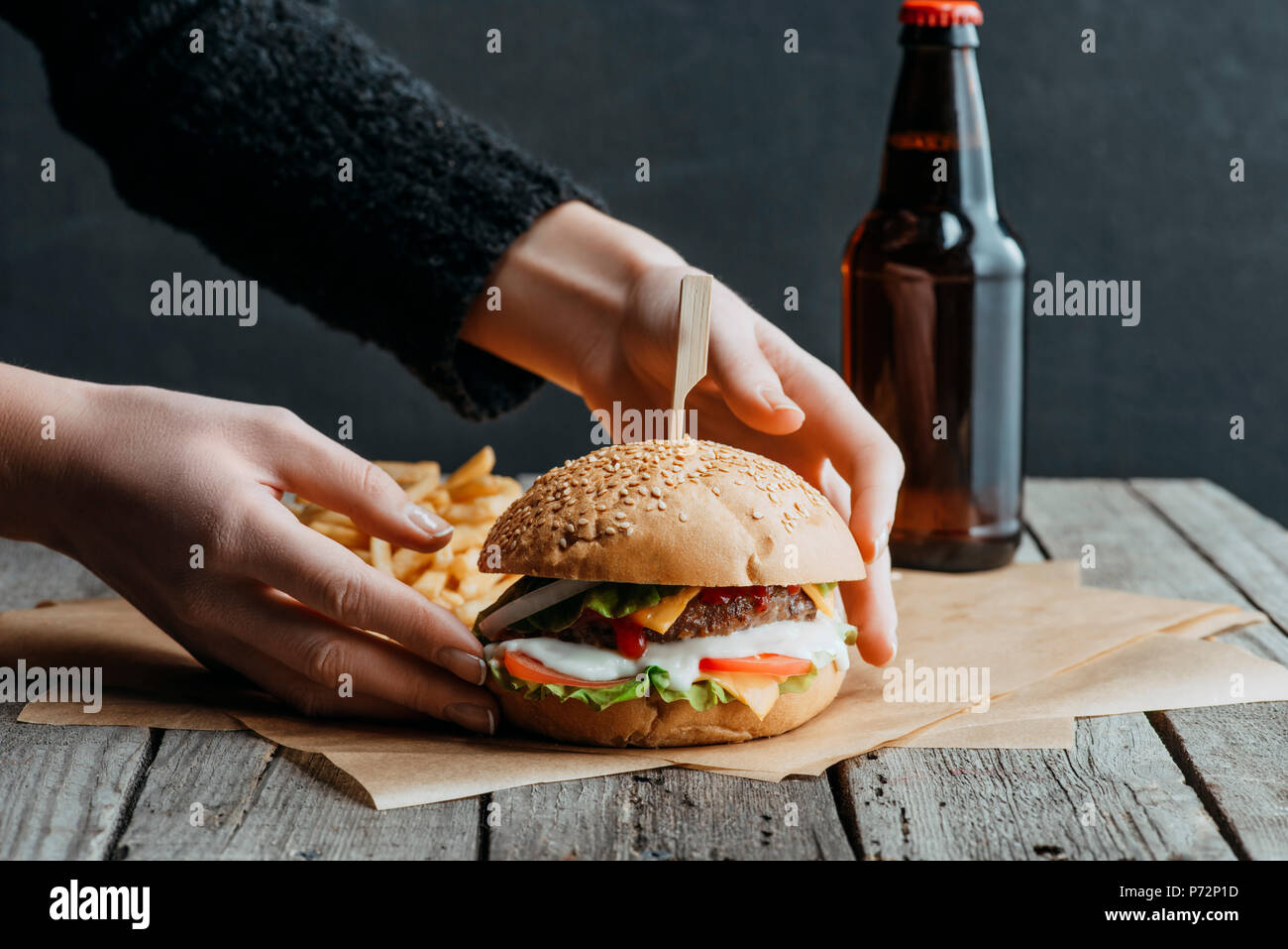 Vista ritagliata della mani femminili con hamburger, patatine fritte e una bottiglia di birra sulla carta da forno sul tavolo di legno Foto Stock