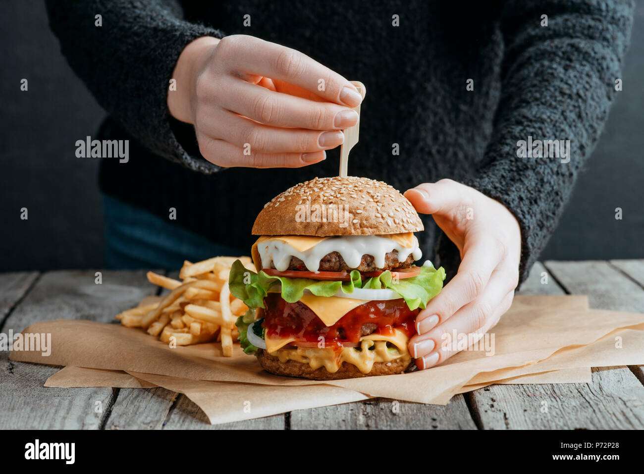 Vista ritagliata della mani femminili con grande gustoso cheeseburger e patatine fritte sulla carta da forno Foto Stock