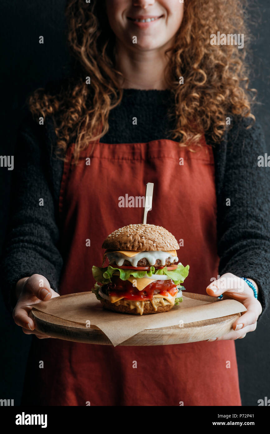 Vista ritagliata della ragazza nel grembiule azienda tagliere in legno con grande in casa cheeseburger Foto Stock