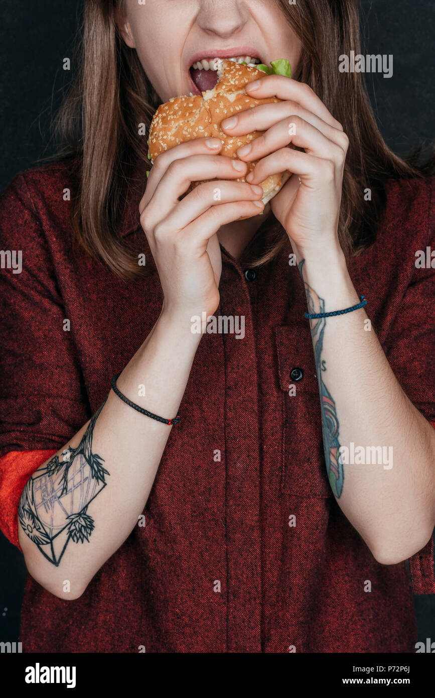 Vista ritagliata della ragazza tatuata mordere gustoso hamburger Foto Stock