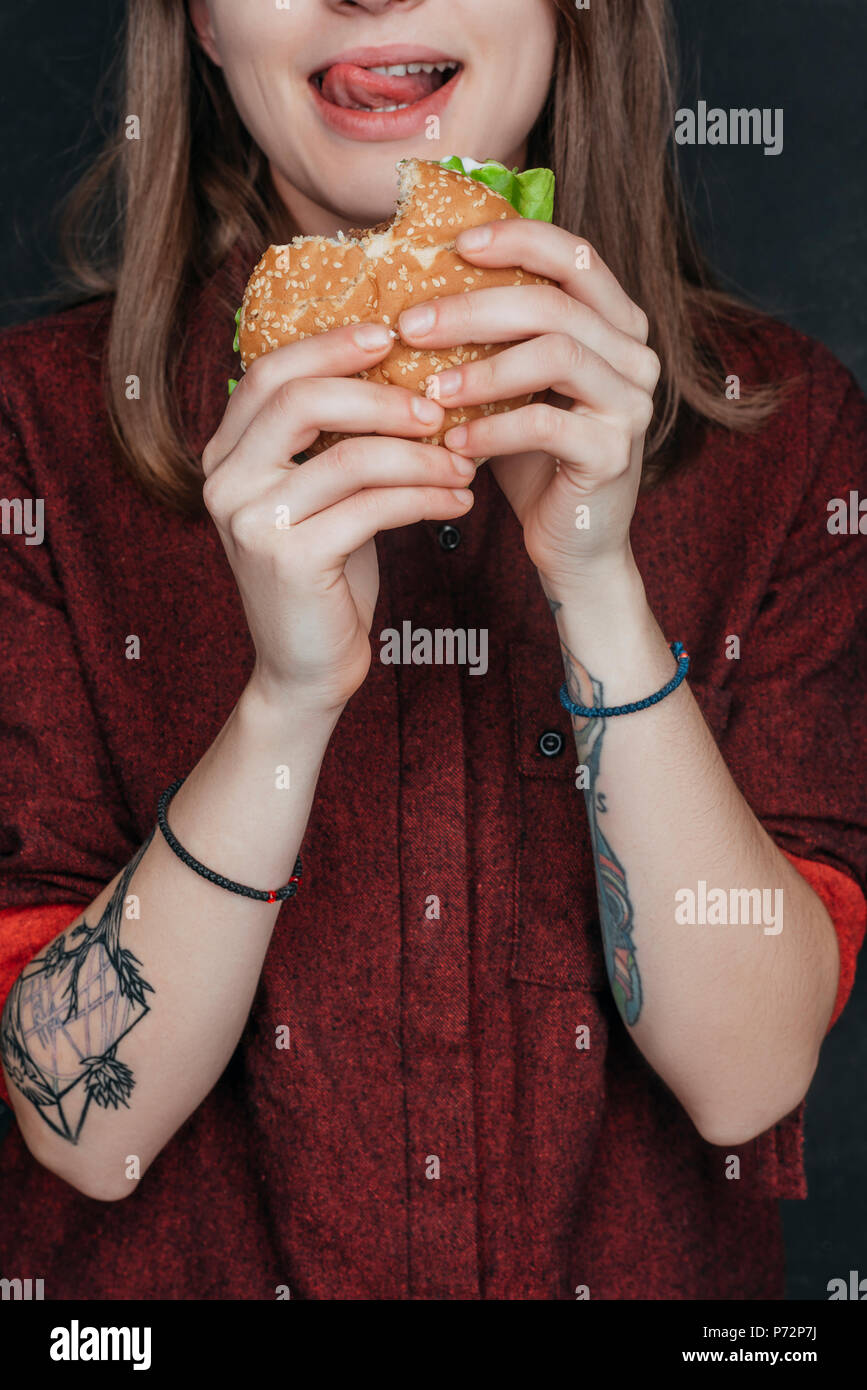 Vista ritagliata della ragazza tatuata mangiare hamburger gustoso Foto Stock