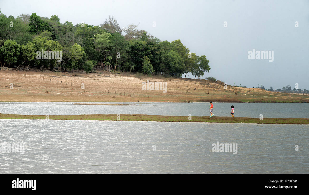 Un ragazzo e una bambina camminando su uno stretto sentiero in Kabini river, Karnataka, India, acqua su entrambi i lati, montagnola e gli alberi in background Foto Stock