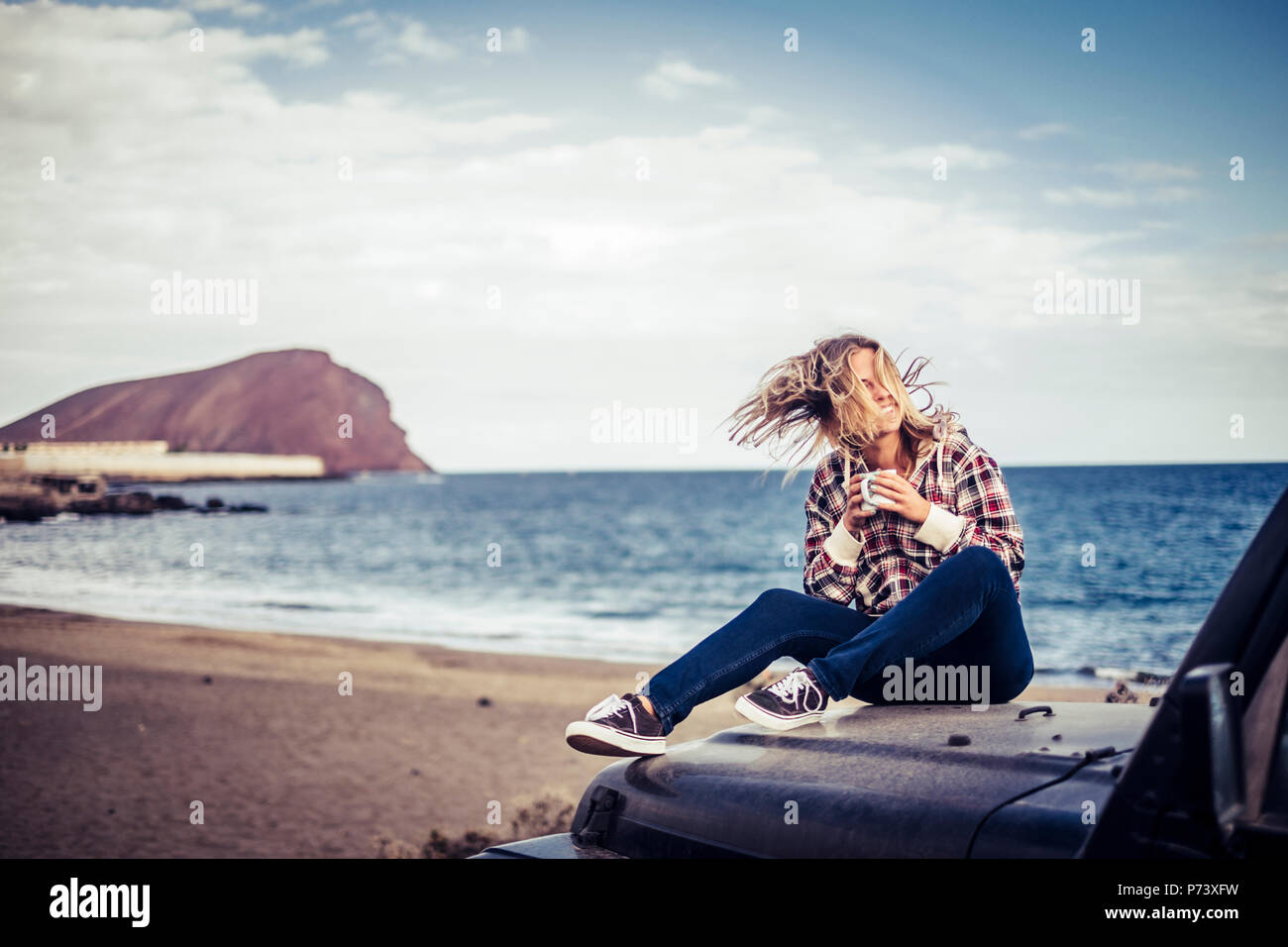 Felice bella bionda ragazza giovane soggiorno all'aperto godendo il tempo e la corsa con off road nero auto. wanderlust concetto parcheggiato sulla sabbia a t Foto Stock