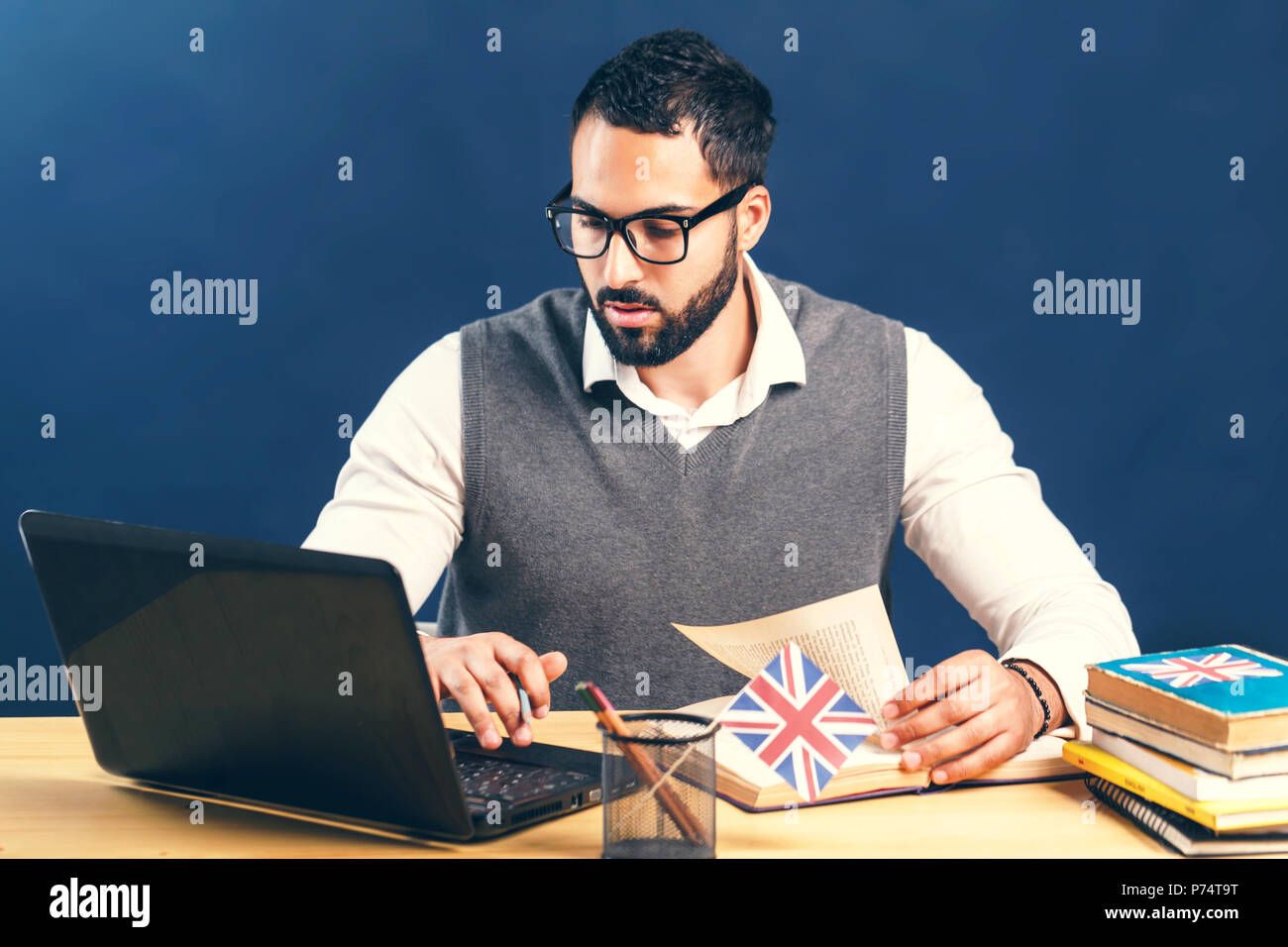 Nero-uomo dai capelli di apprendimento della lingua inglese, che indossa un maglione grigio vest incontaminate e camicia bianca, lavorando sodo alla scrivania da ufficio con computer portatile prima di muro nero Foto Stock