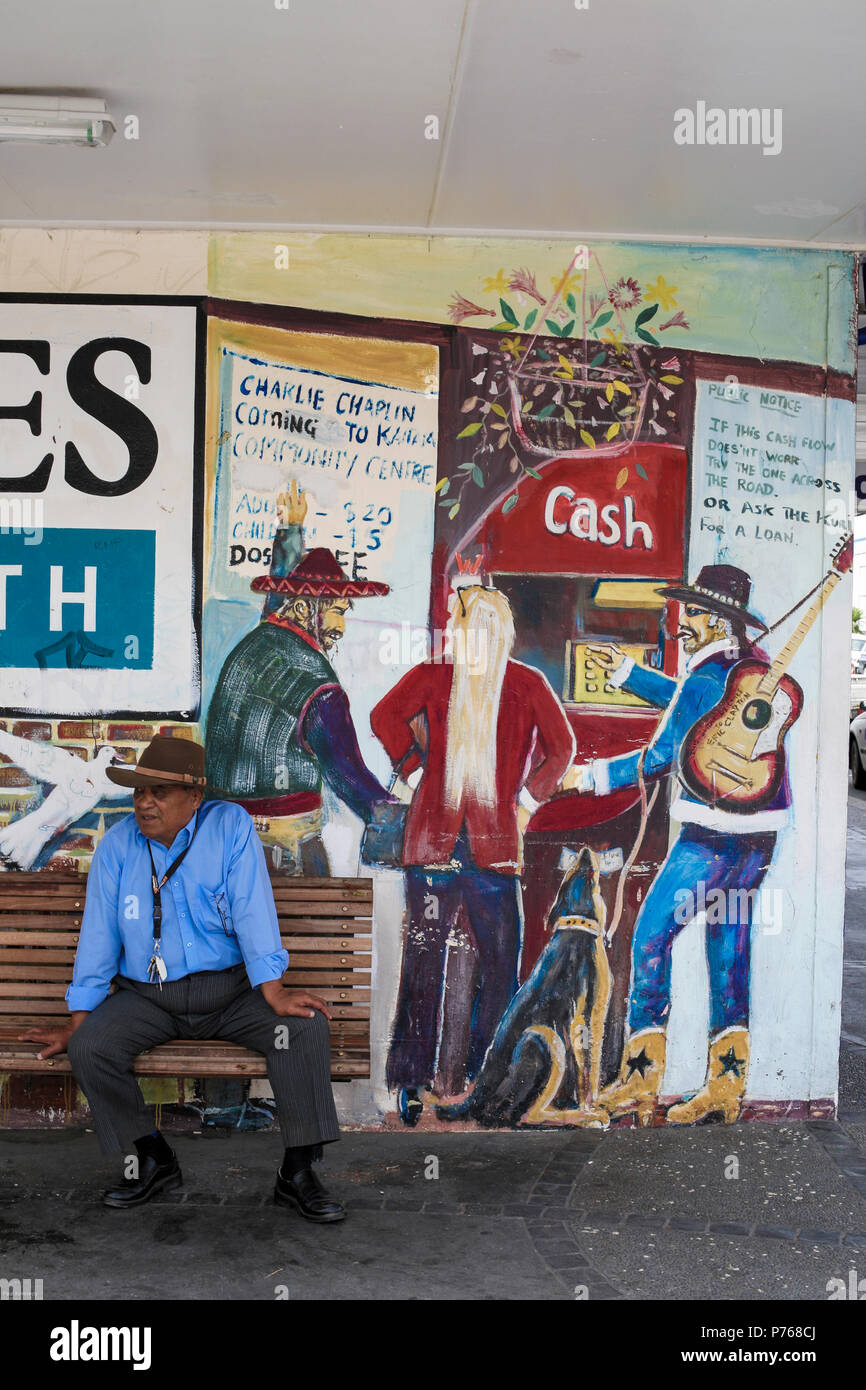 Maori uomo seduto su panchina accanto al murale a Kaitaia, Nuova Zelanda Foto Stock