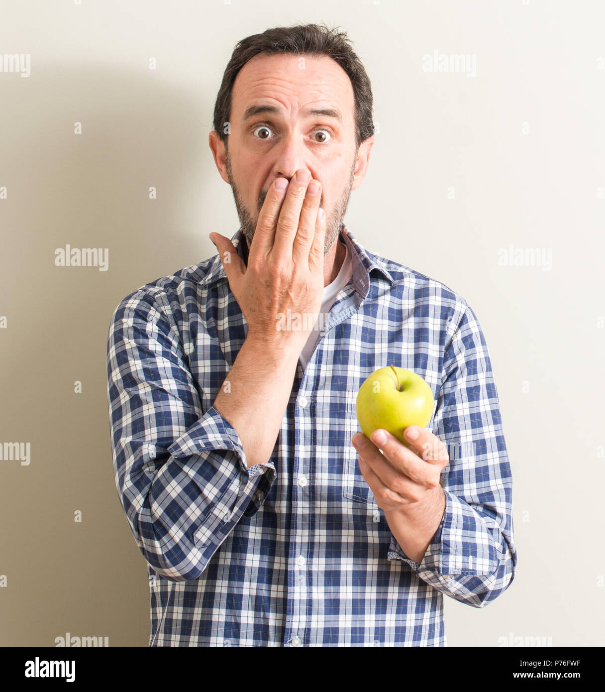 Senior man tenendo una mela verde coprono la bocca con la mano sconvolto con vergogna per errore, espressione di paura, paura in silenzio, concetto segreta Foto Stock