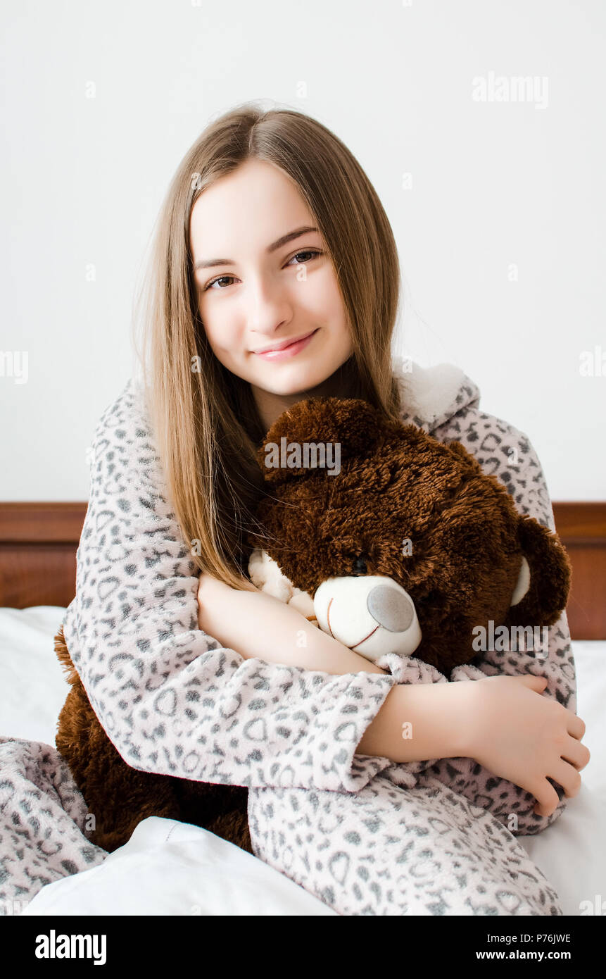 Felice giovane donna con capelli biondi seduta sul letto nella sua camera da letto in un abbraccio con un peluche giocattolo. Marrone preferiti orsacchiotto nelle mani o Foto Stock