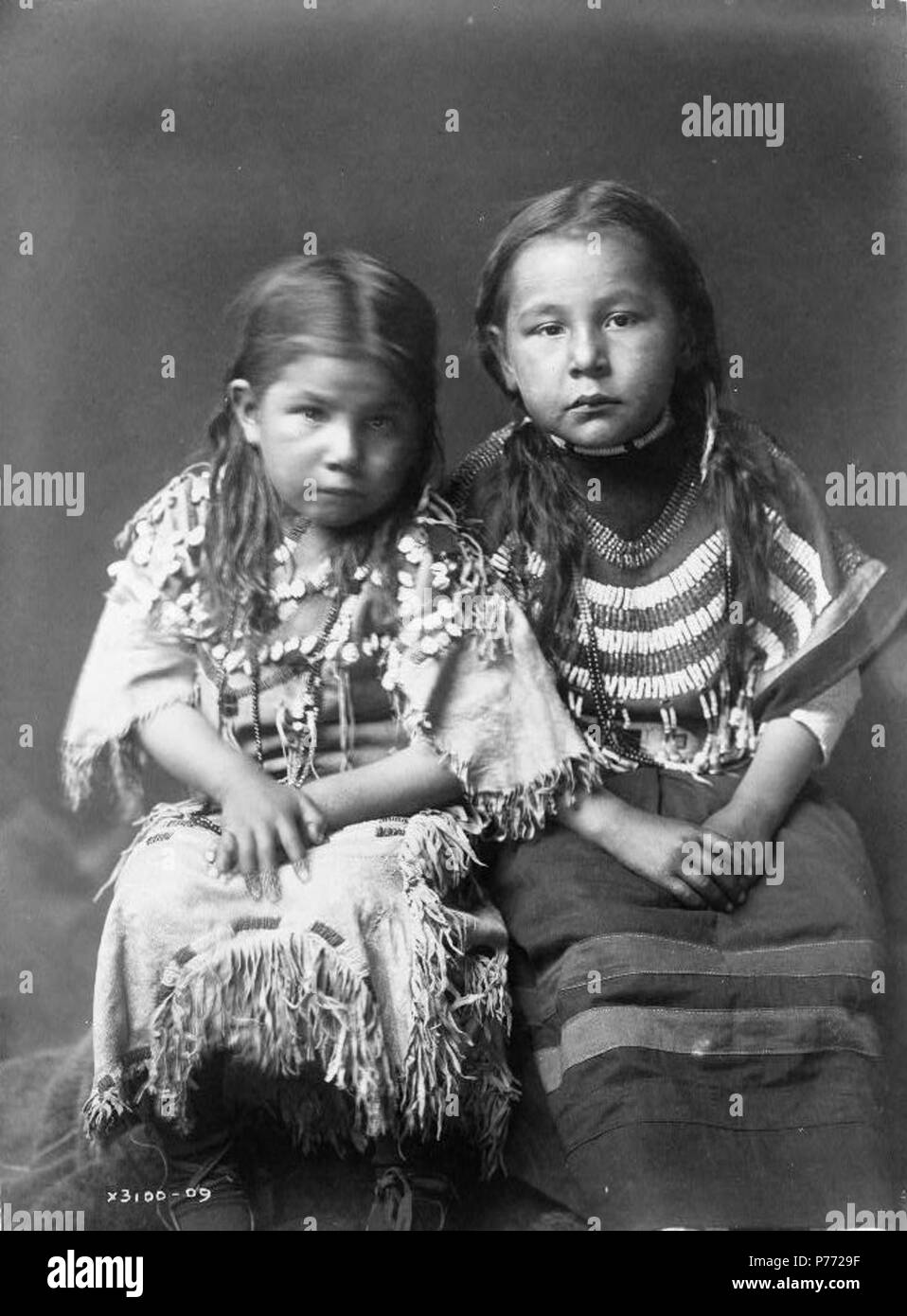 Due ragazze di intera lunghezza, seduto, rivolto verso la parte anteriore, con capelli sciolti, indossando un abito di daino decorate con elks' denti, indossando uno panno bordato abito. circa 1910. 2 Bull scarpa bambini Foto Stock