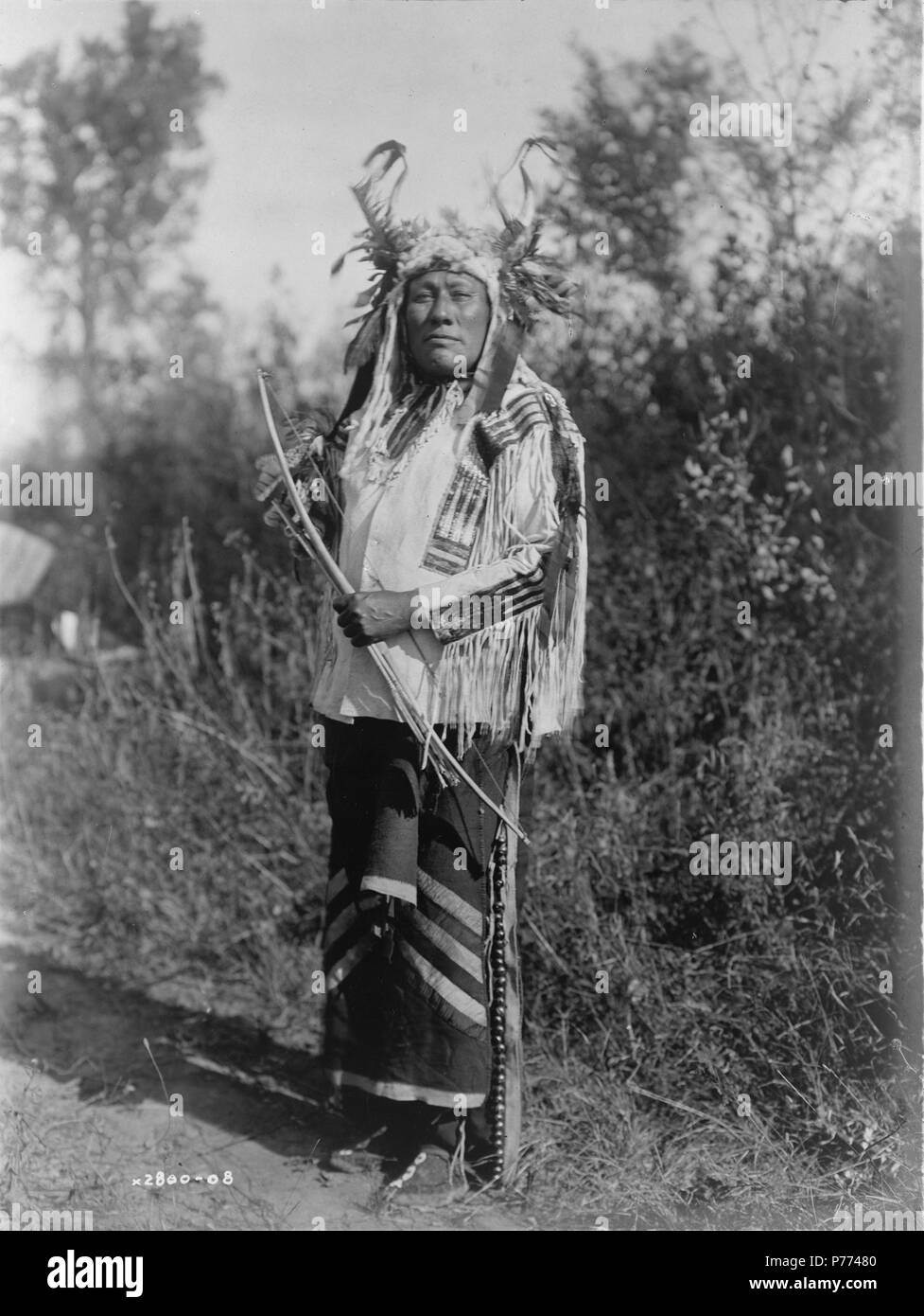 Inglese: Lungo tempo cane, ritratto a figura intera, in piedi, rivolto verso la parte anteriore, indossando headress, maglietta daino e leggings con campane di ottone. circa 1908 8 lungo tempo Hidatsa cane Foto Stock