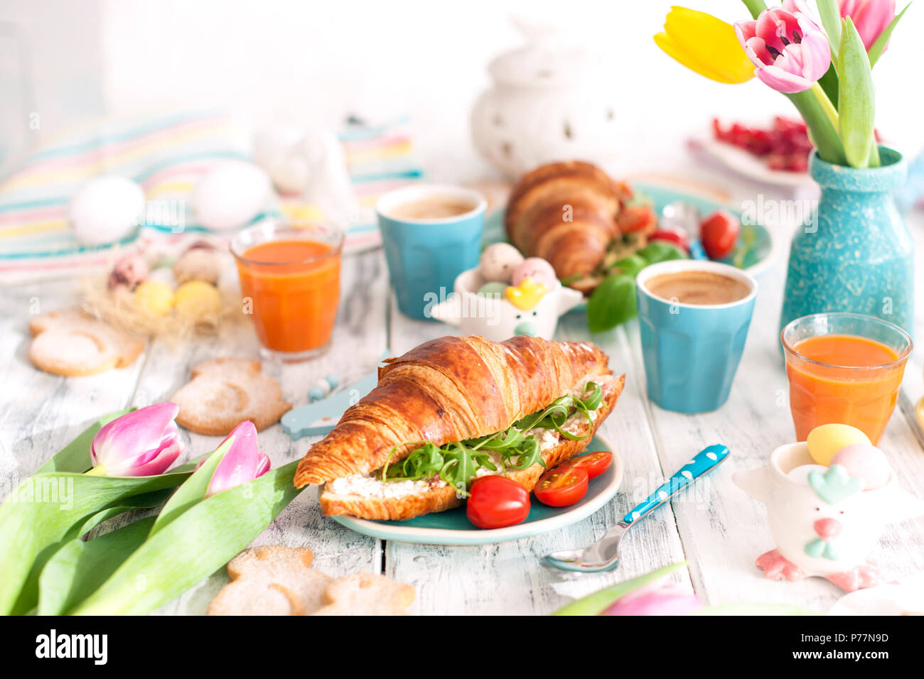 Sfondo con colori diversi. Un family colazione con croissant con rucola e formaggi e caffè aromatico, uova di differenti colori, luminoso piatti e decorazioni di Pasqua, conigli in ceramica Foto Stock