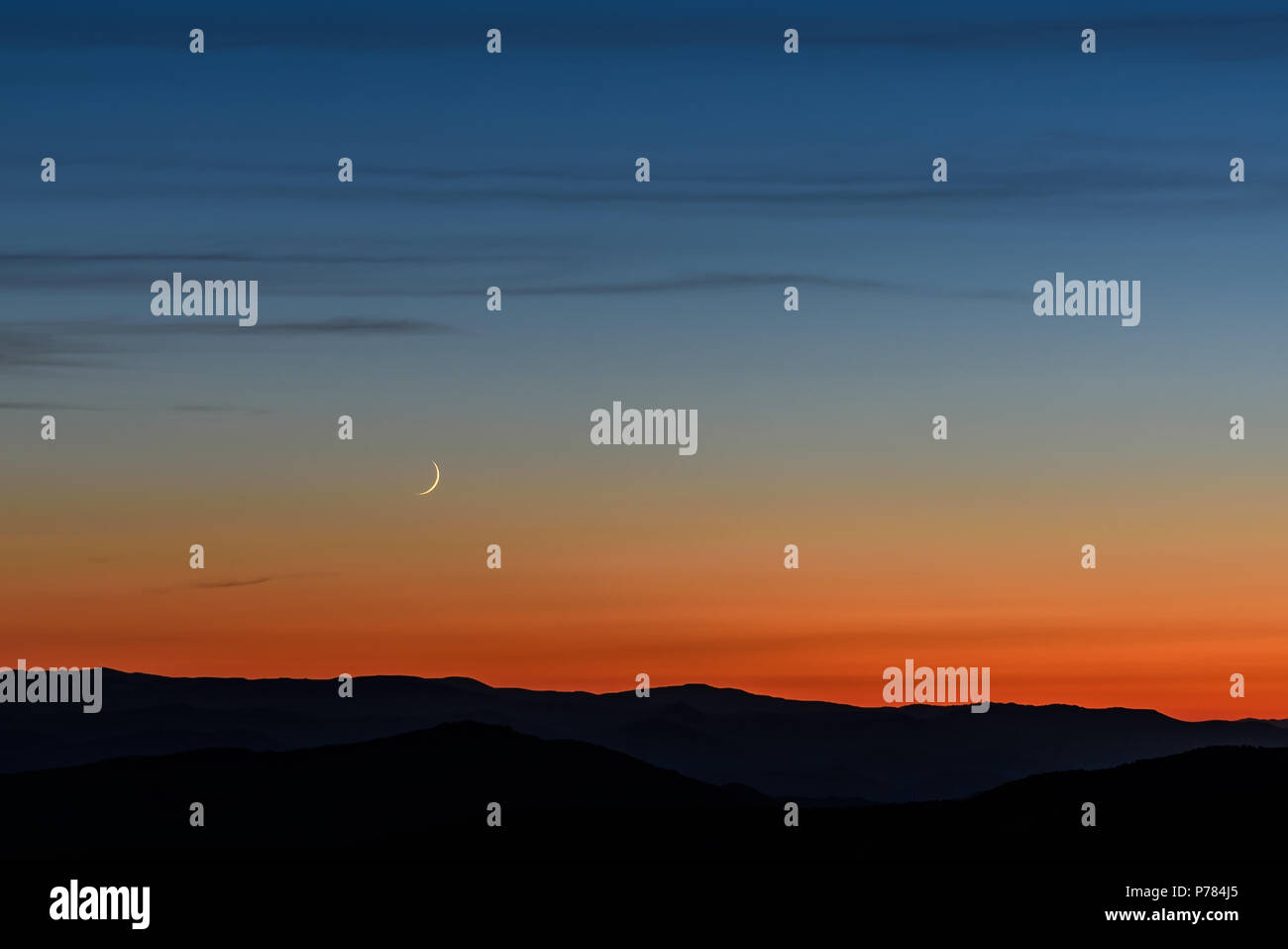 Bella serata paesaggio con una stretta falce di luna e le nuvole nel cielo blu scuro al di sopra delle montagne sullo sfondo di colore arancione sunse Foto Stock