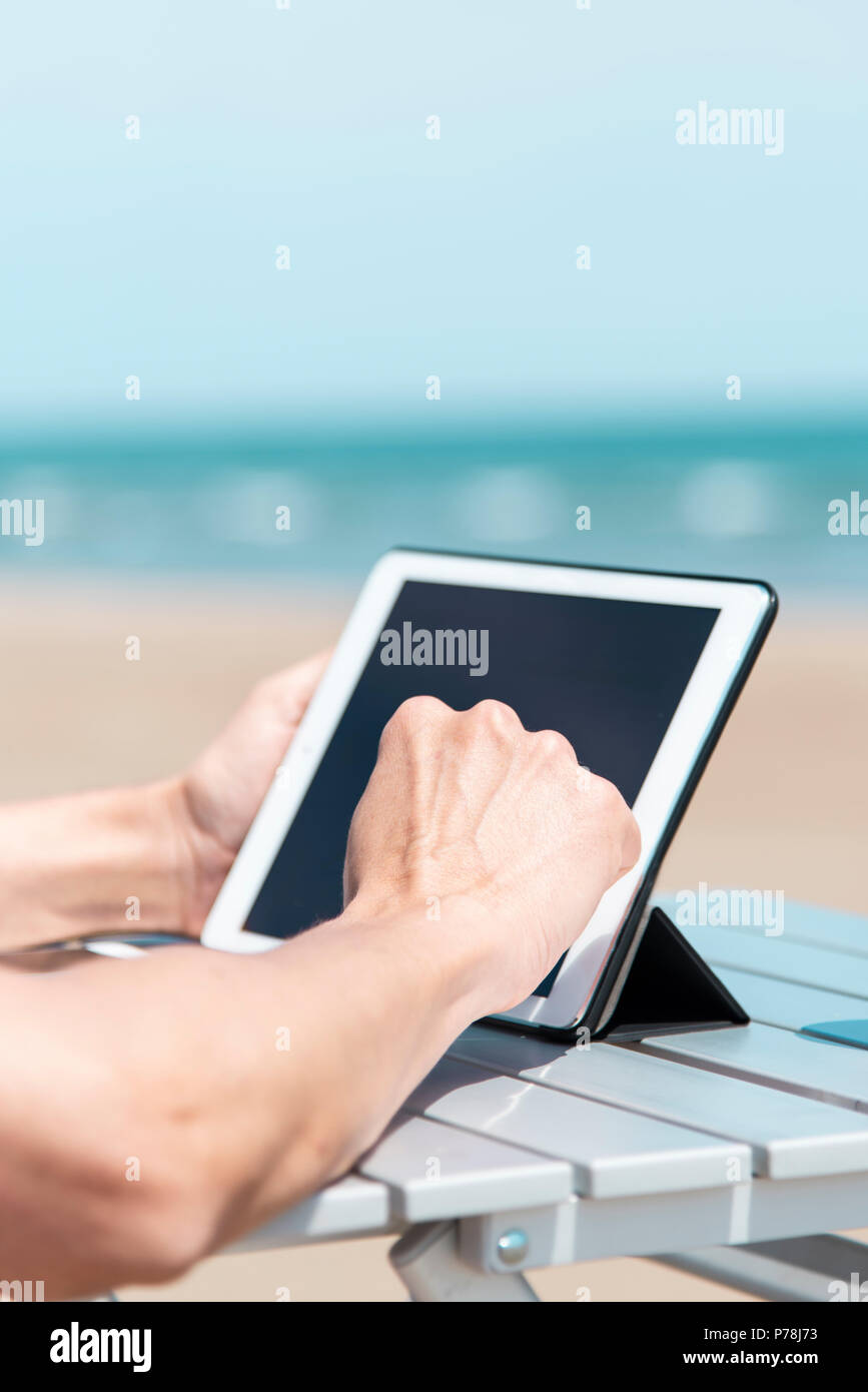 Primo piano di un giovane uomo caucasico sulla spiaggia, utilizzando una tavoletta digitale, seduti a un tavolo all'aperto, con l'oceano sullo sfondo Foto Stock