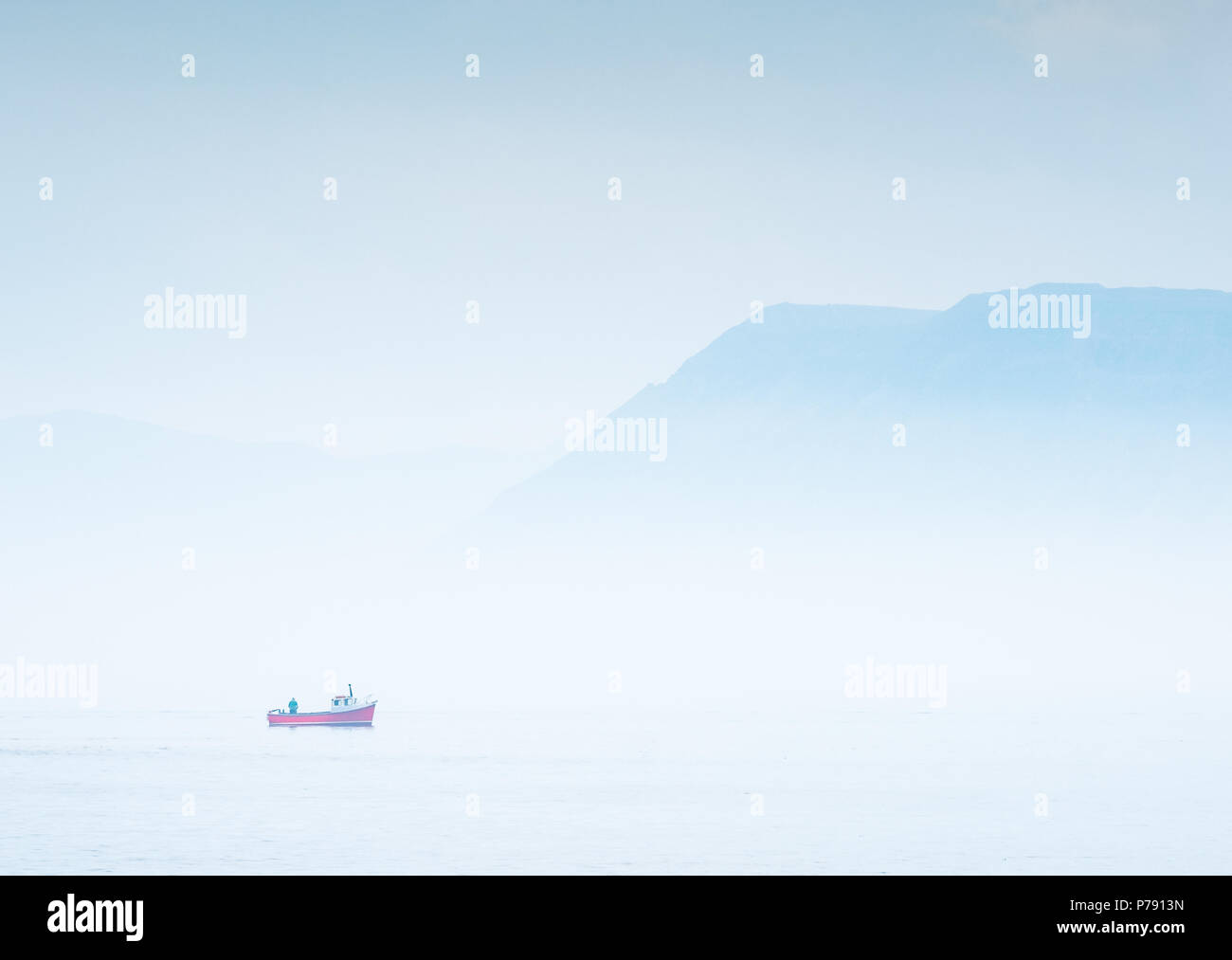 Pescatore solitario e rosso barca da pesca su una tranquilla giornata di nebbia in St George's canale nelle vicinanze Beaumaris sull'Isola di Anglesey nel Galles con sfondo di montagne Foto Stock