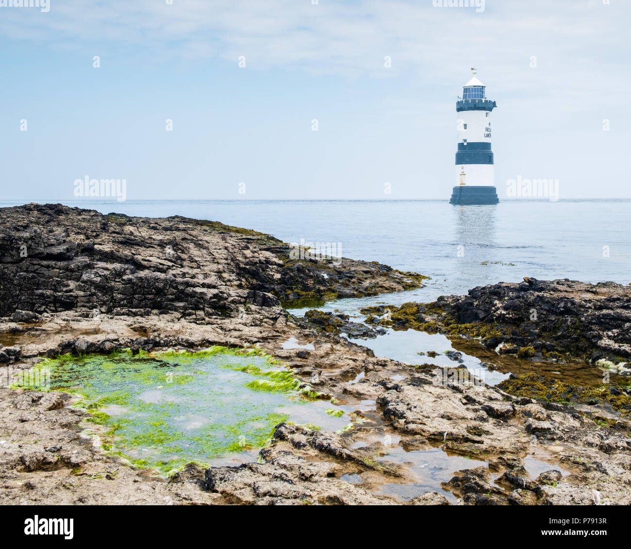 Penmon Lighthouse vicino a Beaumaris, Anglesey, su un luminoso, giornata soleggiata con rocce e alghe verdi rock pool in primo piano ed un cielo blu Foto Stock