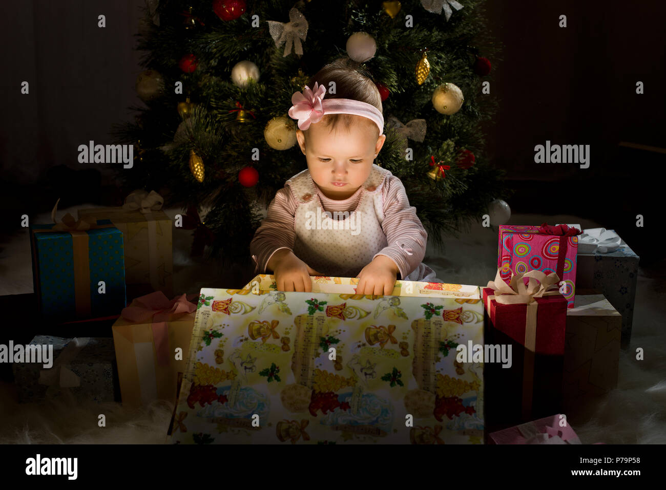 Uno-anno-vecchio bambina solennizzare il Natale, sedersi sotto Natale-tree e raggiungere il dono della scatola, la foto in orizzontale Foto Stock