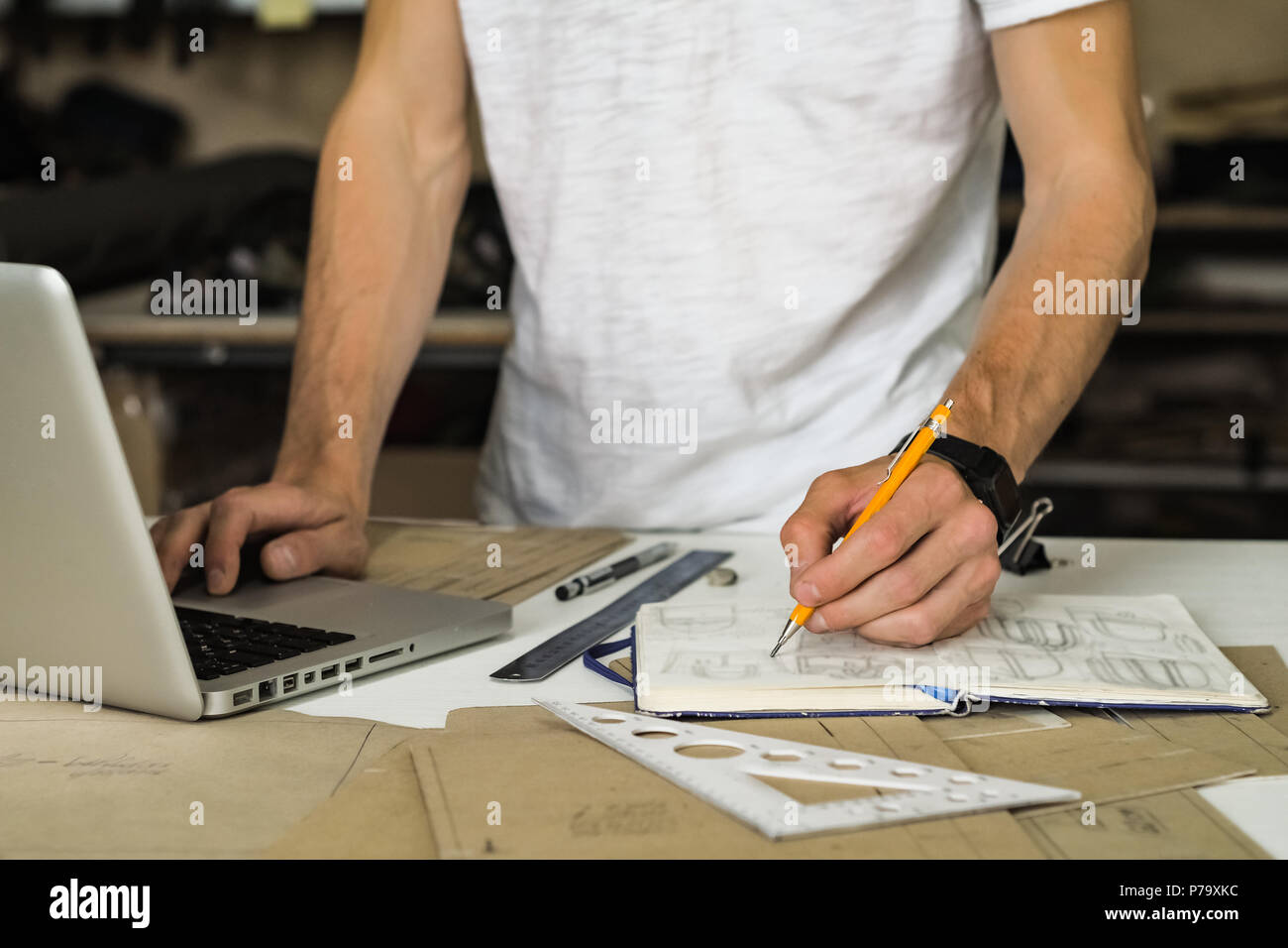 Accessori di consumo o sacchetti designer al lavoro in officina. Le mani del giovane maschio persona con la matita, il progetto di carta e il computer portatile a un tavolo di lavoro Foto Stock