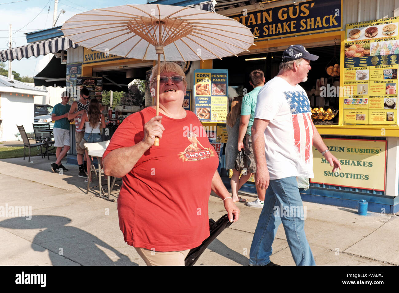 Geneva-On-The-Lake, STATI UNITI D'AMERICA. 4 Luglio, 2018. La donna tiene un ombrellone a temperare il calore estremo mentre si cammina il Geneva-On-The-Lake striscia sulla luglio 4, 2018 vacanza. Il giovane passeggiate passato capitano Gus' Sandwich e agitare Shack, uno dei numerosi stabilimenti di catering per il 4 luglio 2018 la folla in questa cittadina festeggia il noi Giorno di Indipendenza. Contrassegnare Kanning/Alamy Live News Foto Stock