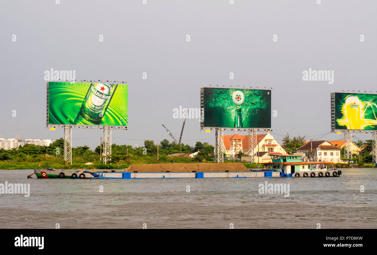 Trasporto alla rinfusa inclus. Il trasporto di un carico sul fiume Saigon con la mitica Heineken cartelloni in background., Ho Chi Minh City, Vietnam. Foto Stock
