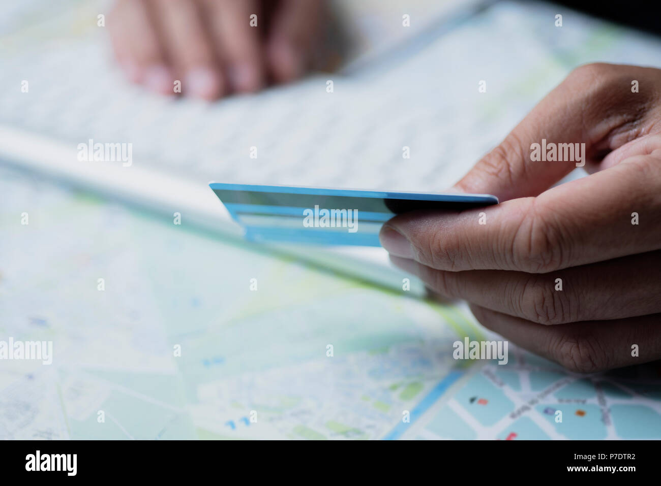 Primo piano di un uomo caucasico utilizzando una carta di credito per prenotare un viaggio online utilizzando il suo computer su un tavolo pieno di mappe Foto Stock