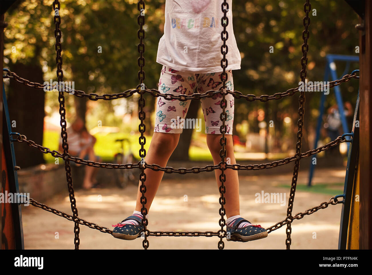 Bambina sul parco giochi nel parco, arrampicate sulle catene, le persone più anziane con un bambino piccolo in sfondo sfocato. Foto Stock